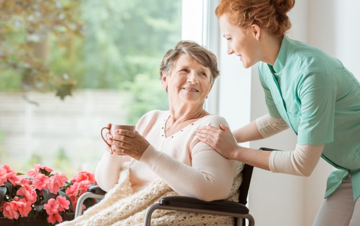 A professional caretaker in uniform helping a geriatric female patient on a wheelchair. Senior holding a cup and sitting by a large window in a rehabilitation center.